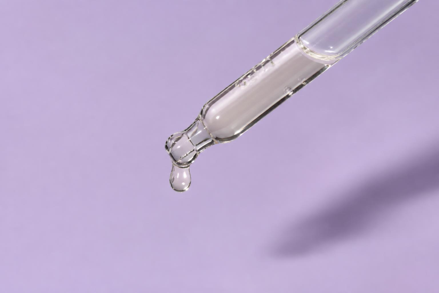 Glass pipette releasing a clear, faintly pale-yellow vitamin C serum drop against a lilac background